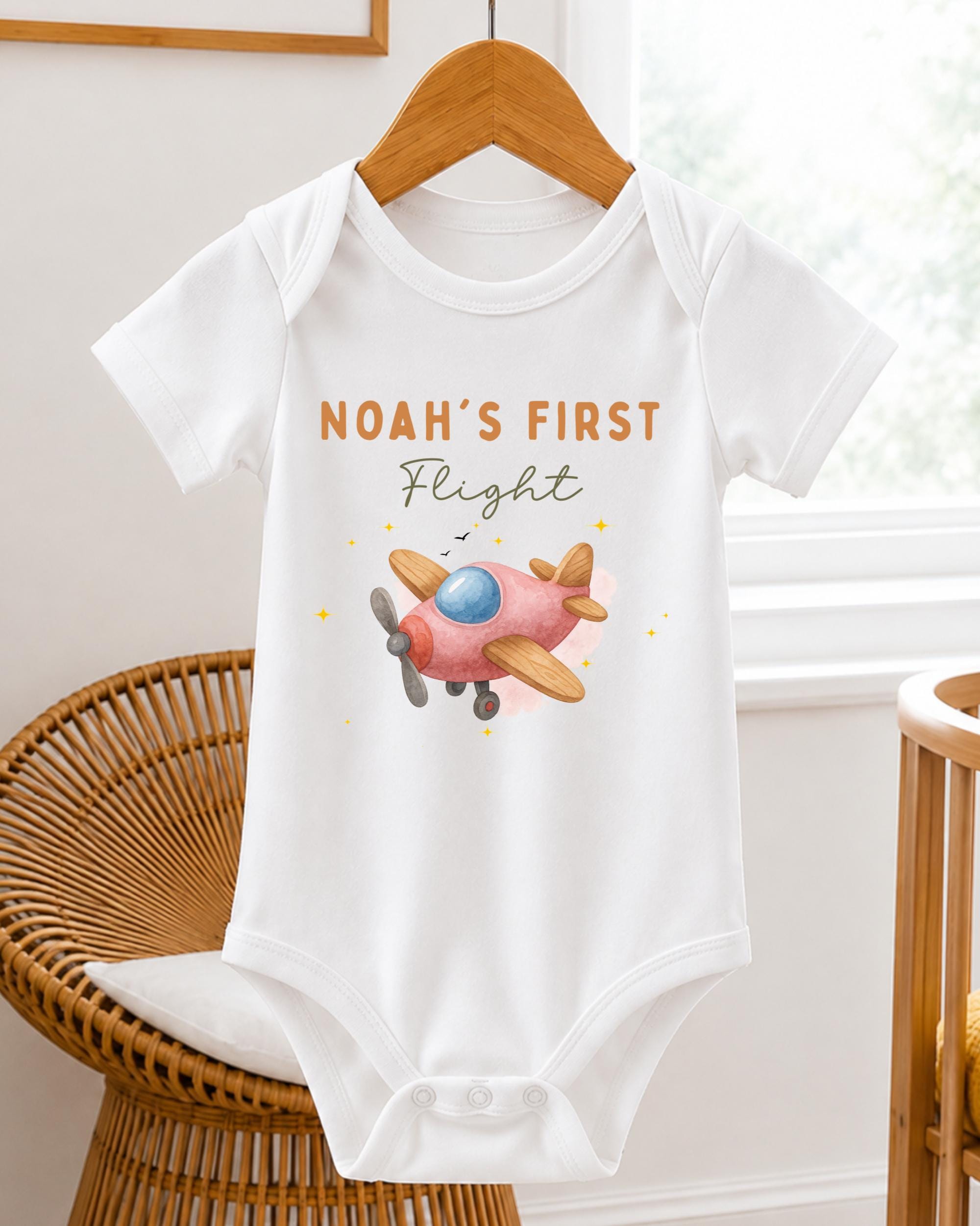 A white baby onesie with a pink airplane printed on it, hanging on a wooden hanger.