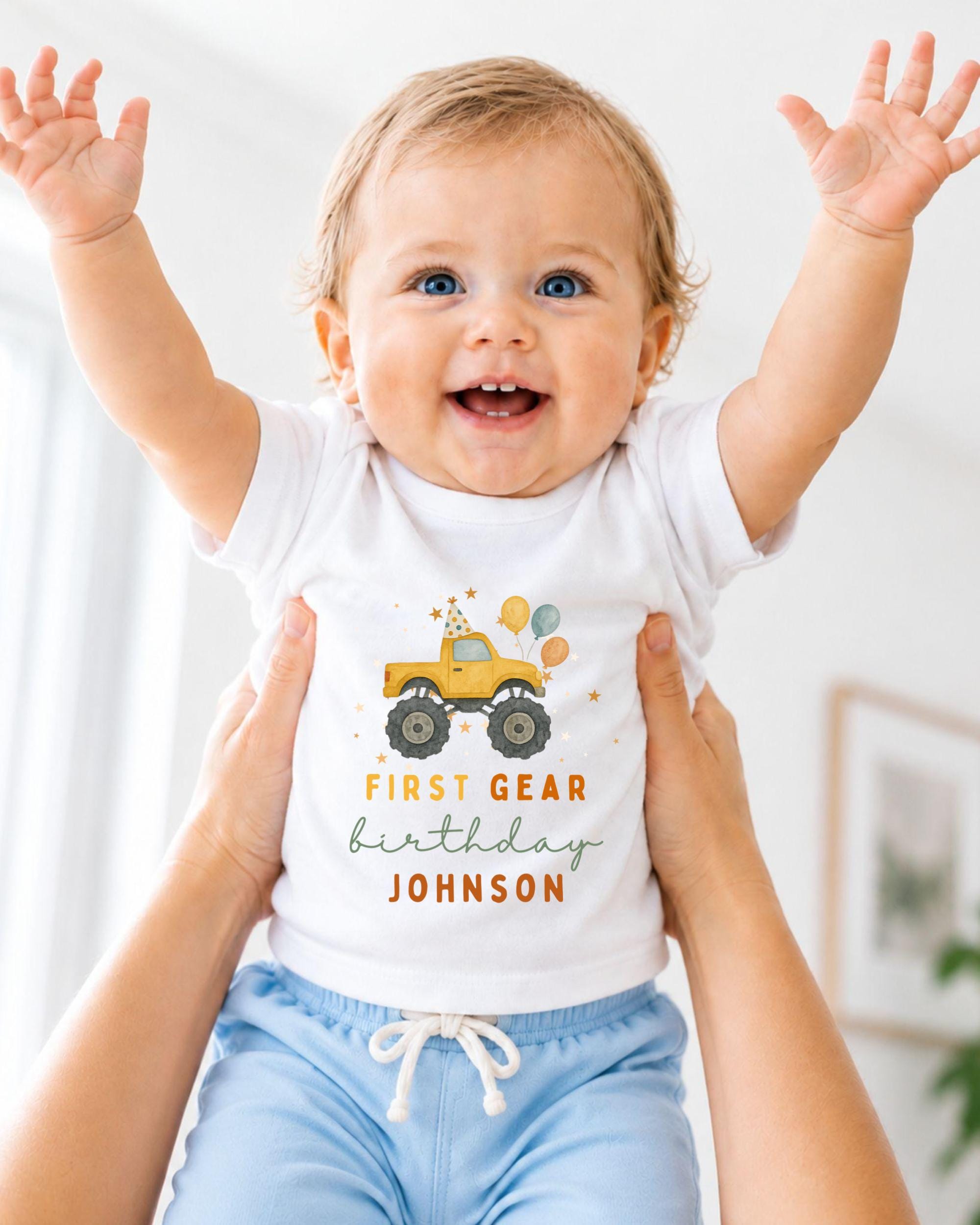 A smiling baby wearing a white t-shirt with a first birthday truck graphic and the text "FIRST GEAR birthday JOHNSON" is being held up by two people.