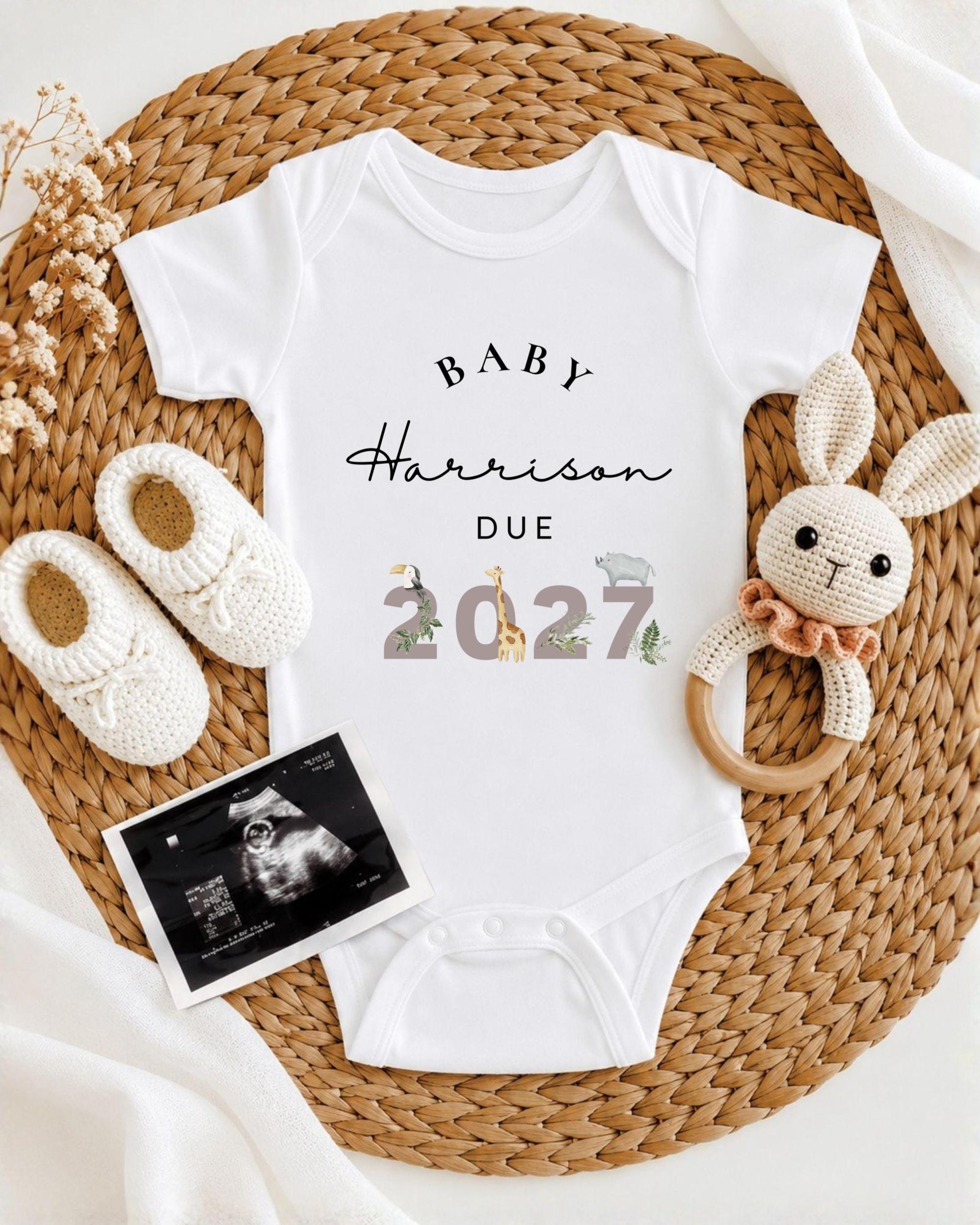 A baby's onesie with the text "BABY Harrison Due 2027" printed on it, along with a crocheted rabbit toy and a pair of white baby shoes.
