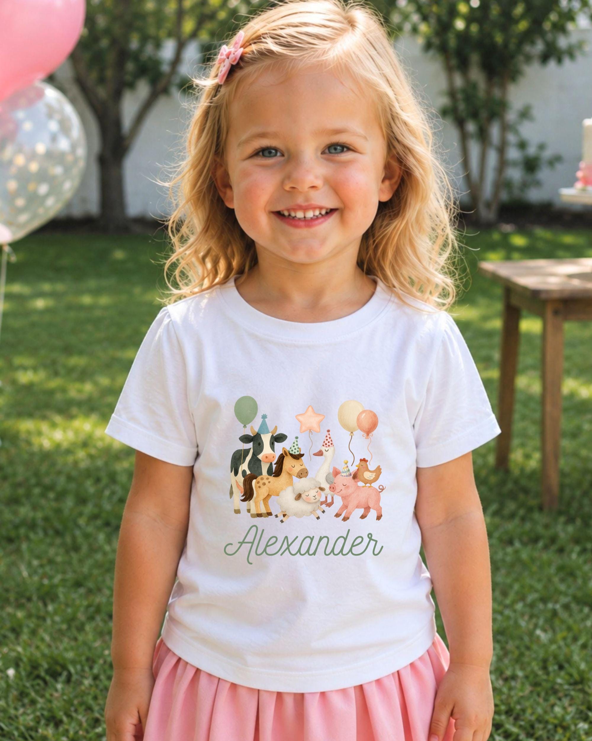 A young girl with blonde hair is smiling and wearing a white t-shirt with a graphic of animals and balloons, standing in a grassy area with trees in the background.