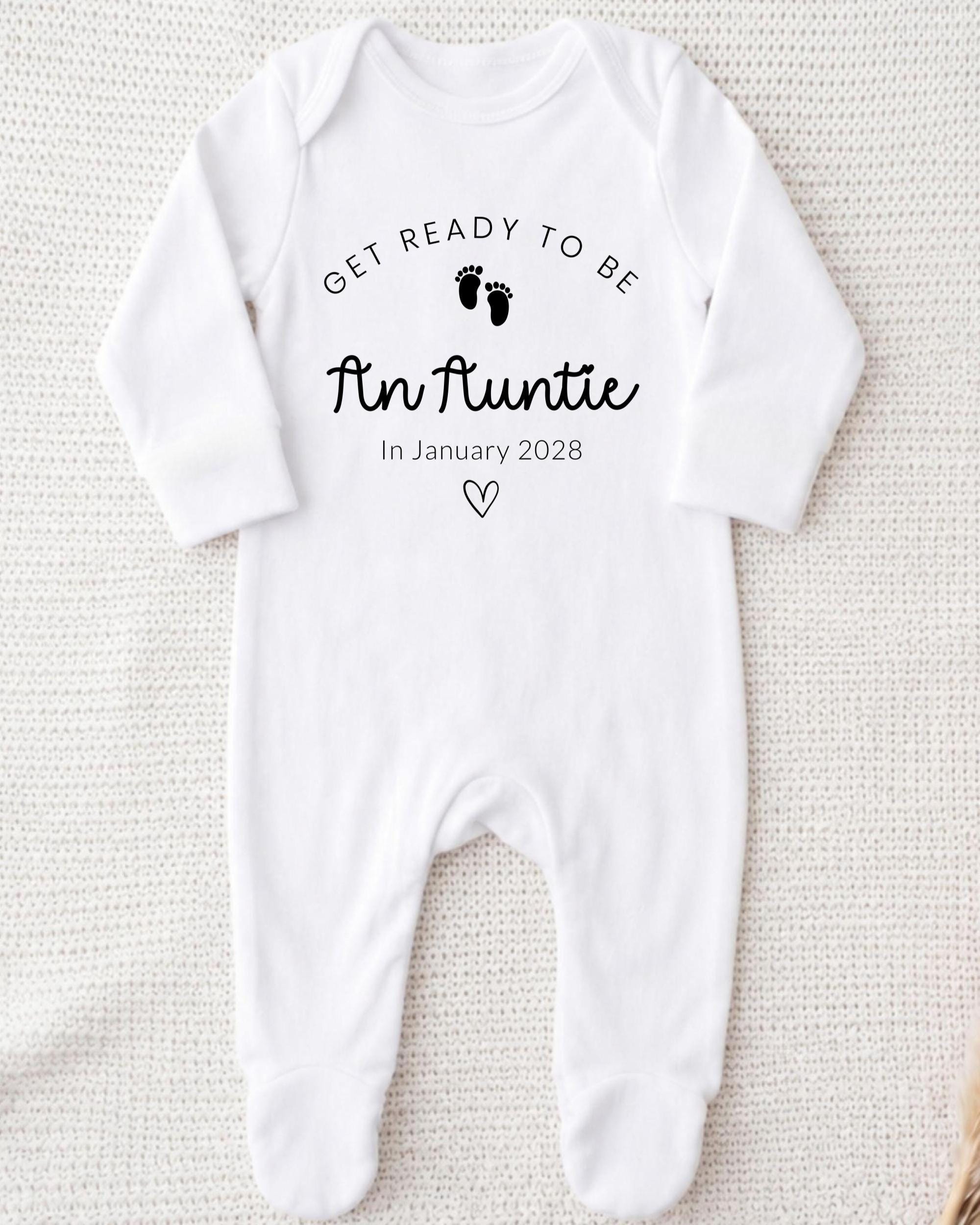 A white onesie with the text "Get ready to be An Auntie" printed on it, along with a heart symbol and the date "In January 2028".