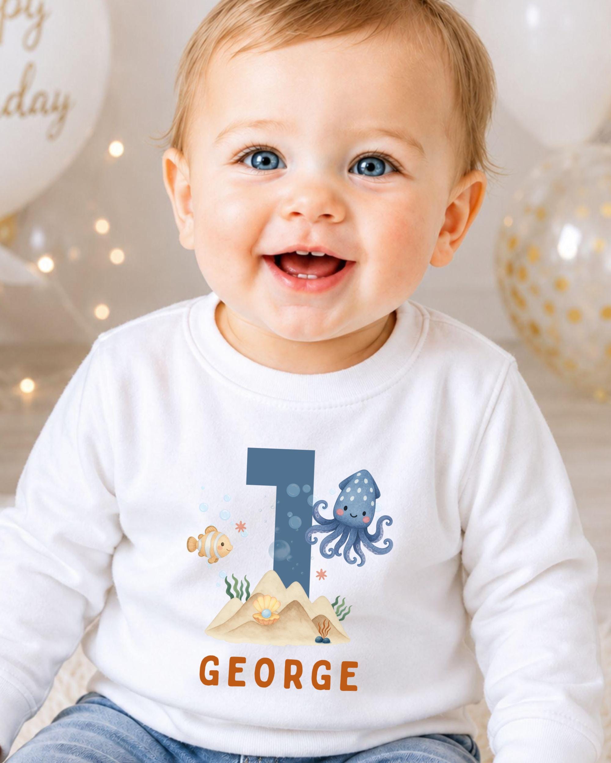 A smiling baby boy wearing a white shirt with a blue number 1 and an illustration of an octopus and a fish.