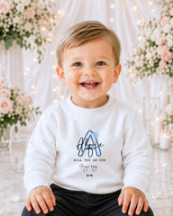 A smiling baby boy wearing a white sweater with a blue logo and text, sitting in front of a floral background with white lights.
