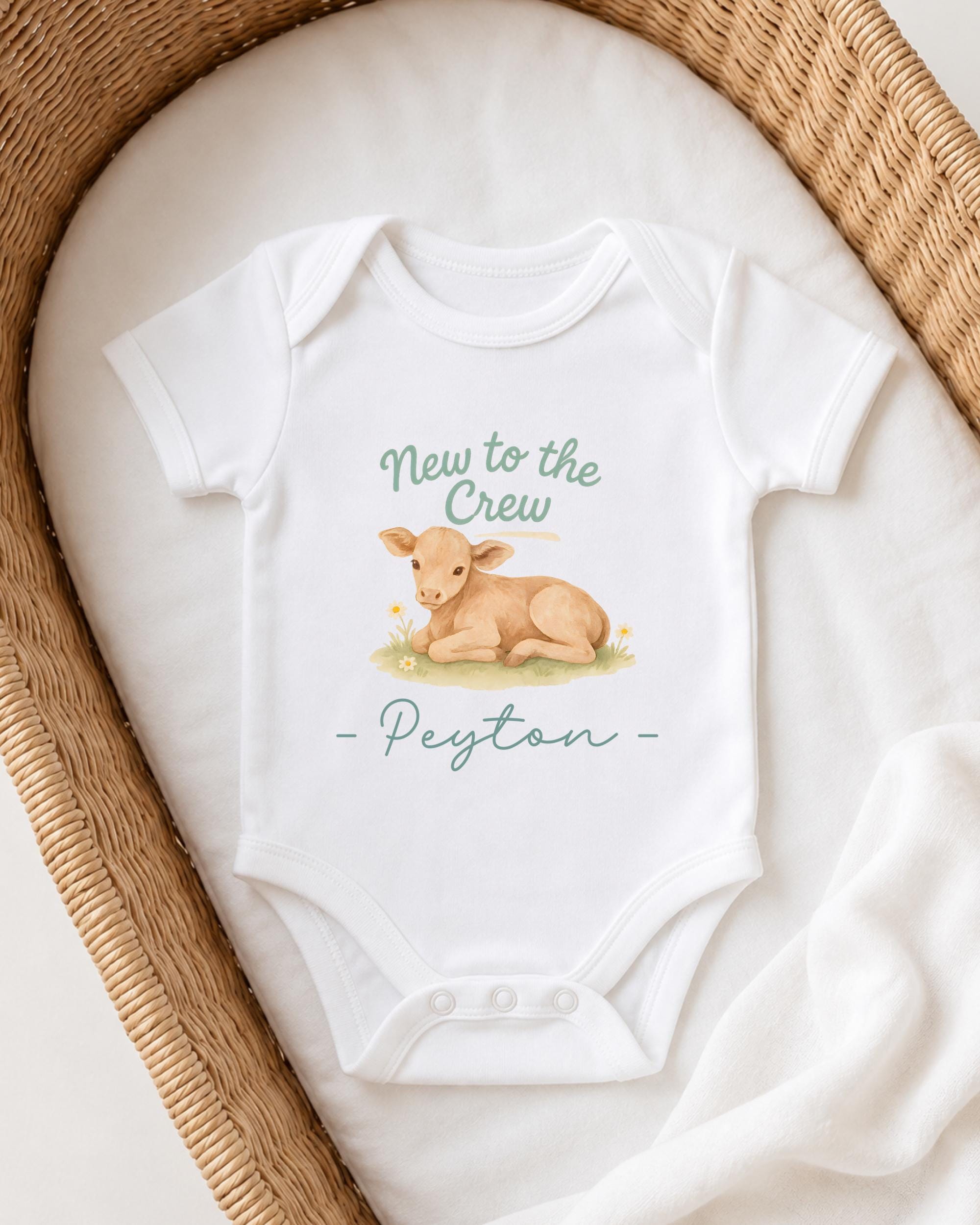 A white baby onesie with a printed image of a baby lamb and the text "New to the Crew Peyton" printed on it, placed on a white blanket.