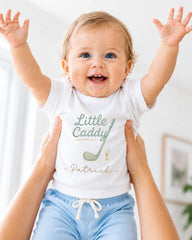 A smiling baby wearing a white t-shirt with a golf club and the name "Patrick" printed on it, being held up by someone's arms.