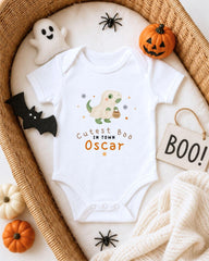 A white onesie with a cartoon dinosaur and the text "Cute Boo Oscar" is displayed on a bed, surrounded by Halloween decorations such as a ghost, a pumpkin, a bat, and a sign that says "BOO!".