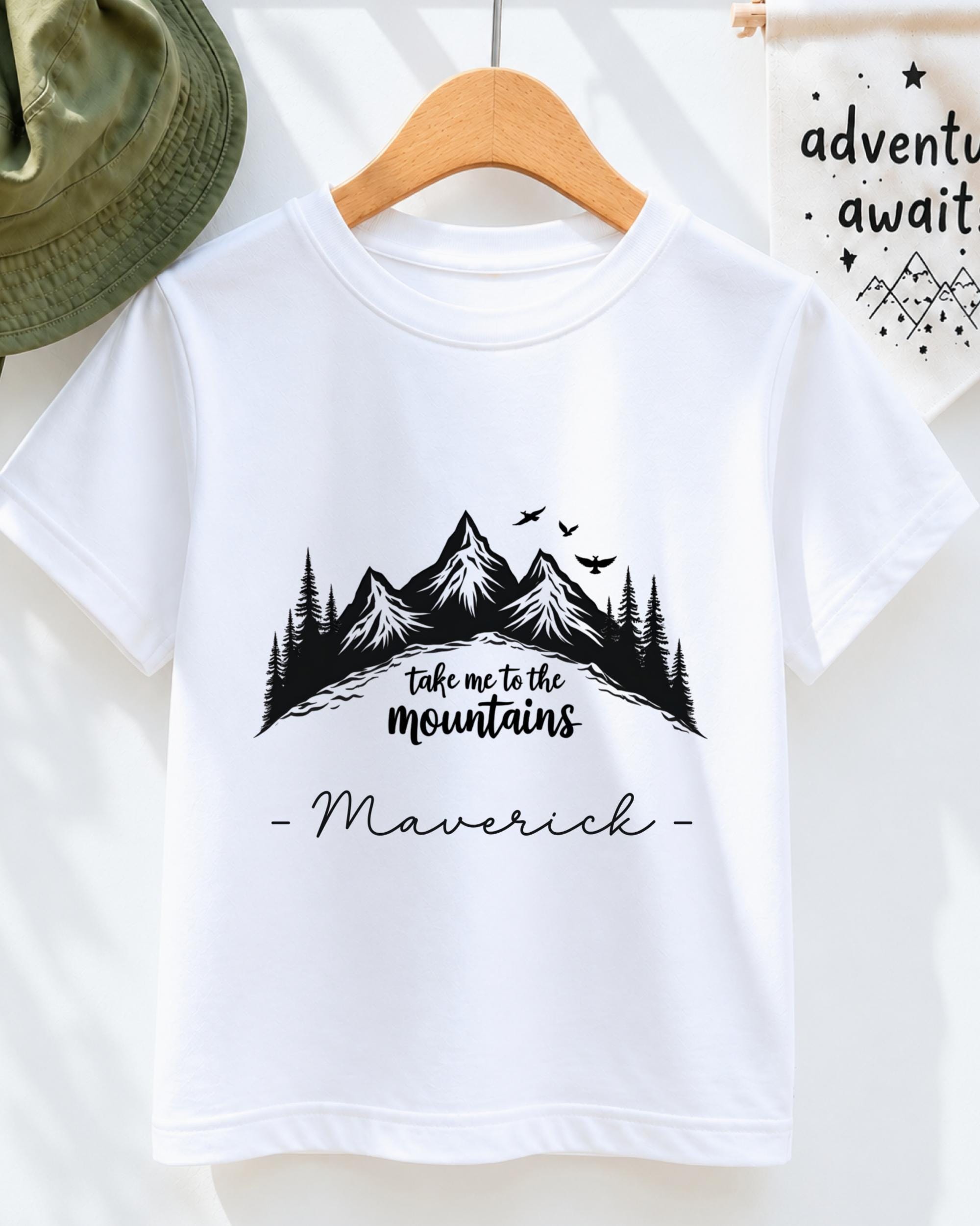 A white t-shirt with a mountain landscape design and the text "Take me to the mountains" printed on it, along with the name "Maverick" below the design.