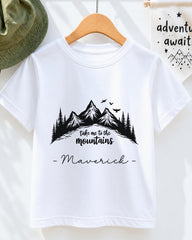 A white t-shirt with a mountain landscape design and the text "Take me to the mountains" printed on it, along with the name "Maverick" below the design.