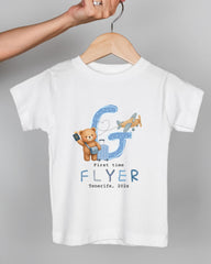 A white t-shirt with a graphic design featuring a teddy bear, a letter "G", and an airplane. The text below the design says "First time FLYER Tenerife, 20x".