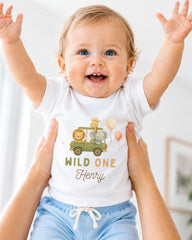 A smiling baby wearing a white t-shirt with a cartoon car and animals, being held up by someone's arms.