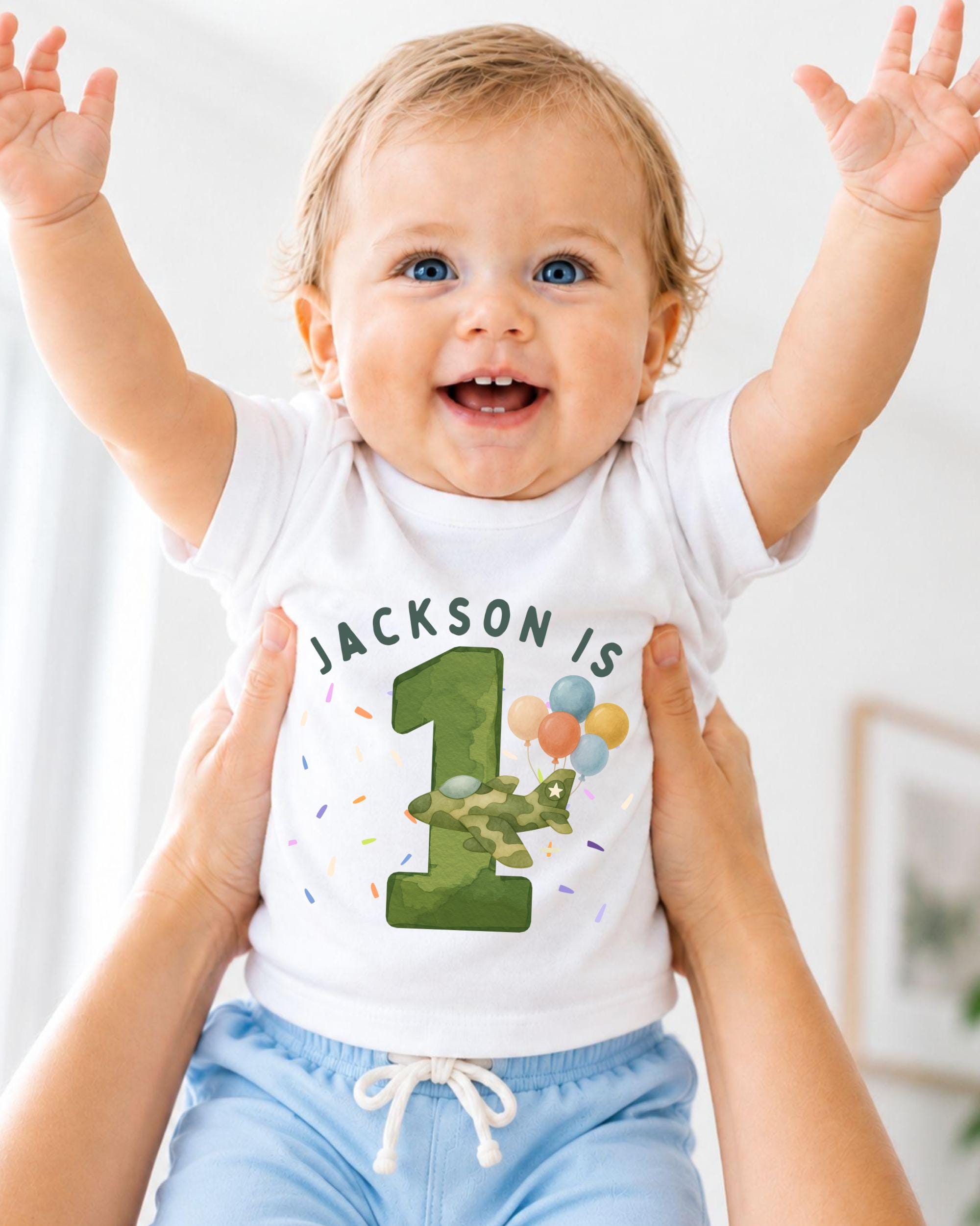 A smiling baby wearing a white t-shirt with the text "JACKSON IS 1" and a graphic of a green airplane with balloons.