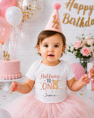A young girl wearing a white t-shirt with a teddy bear and the text "Halfway to 1" is sitting in front of a birthday cake and balloons, with a "Happy Birthday" sign in the background.