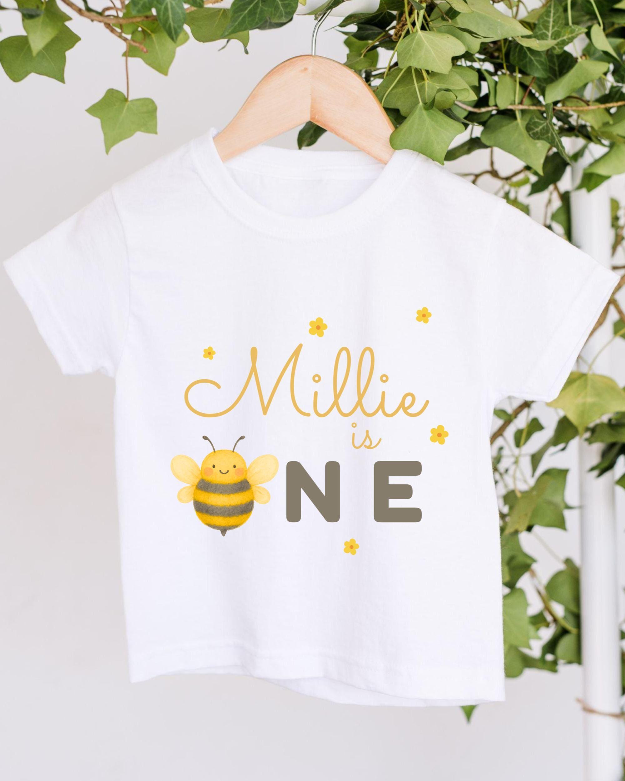 A white t-shirt with a yellow bee graphic and the text "Milly is one" printed on it, hanging on a wooden hanger.