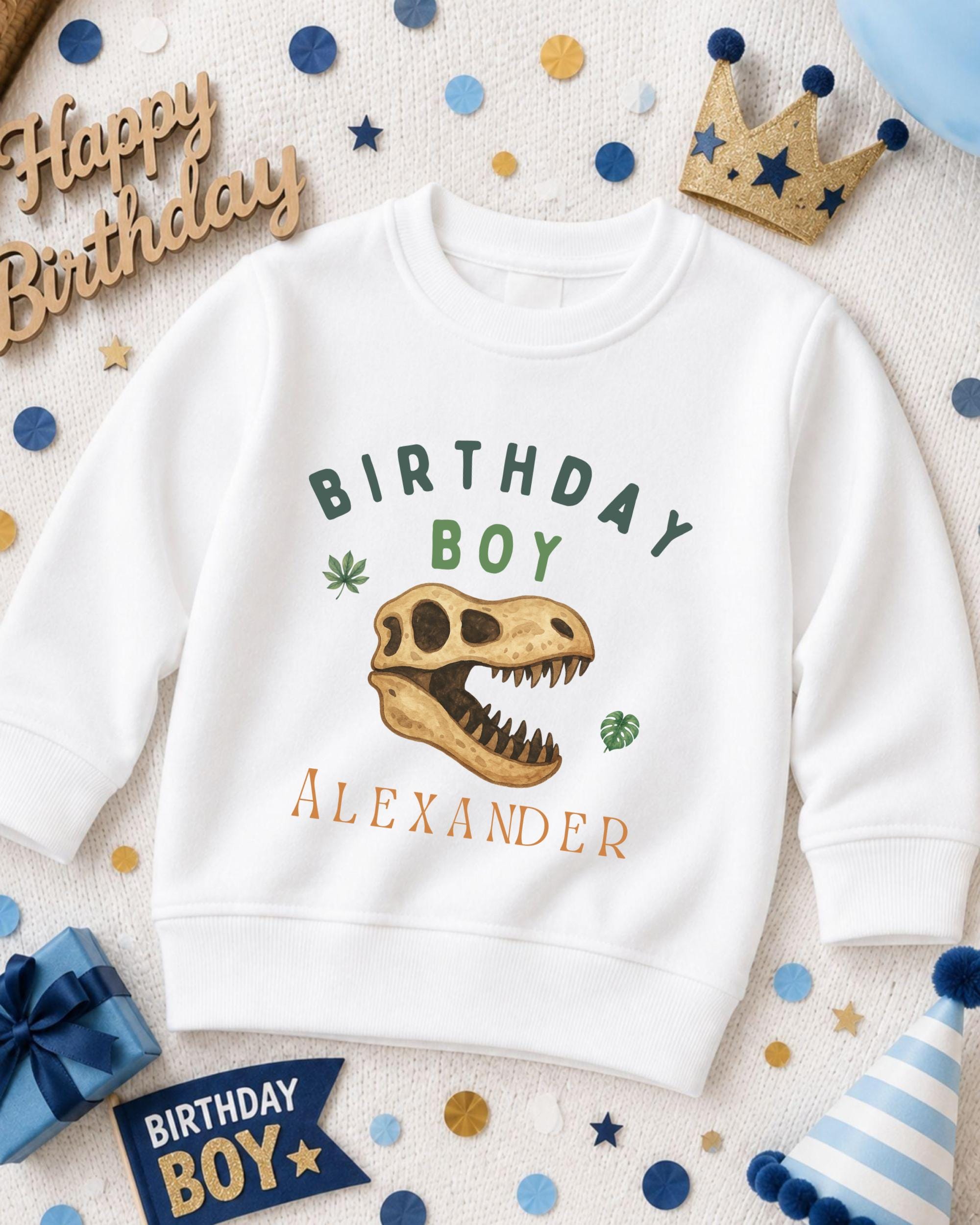 A white sweatshirt with a dinosaur skull design and the text "Birthday Boy Alexander" printed on it, surrounded by birthday decorations and a crown.