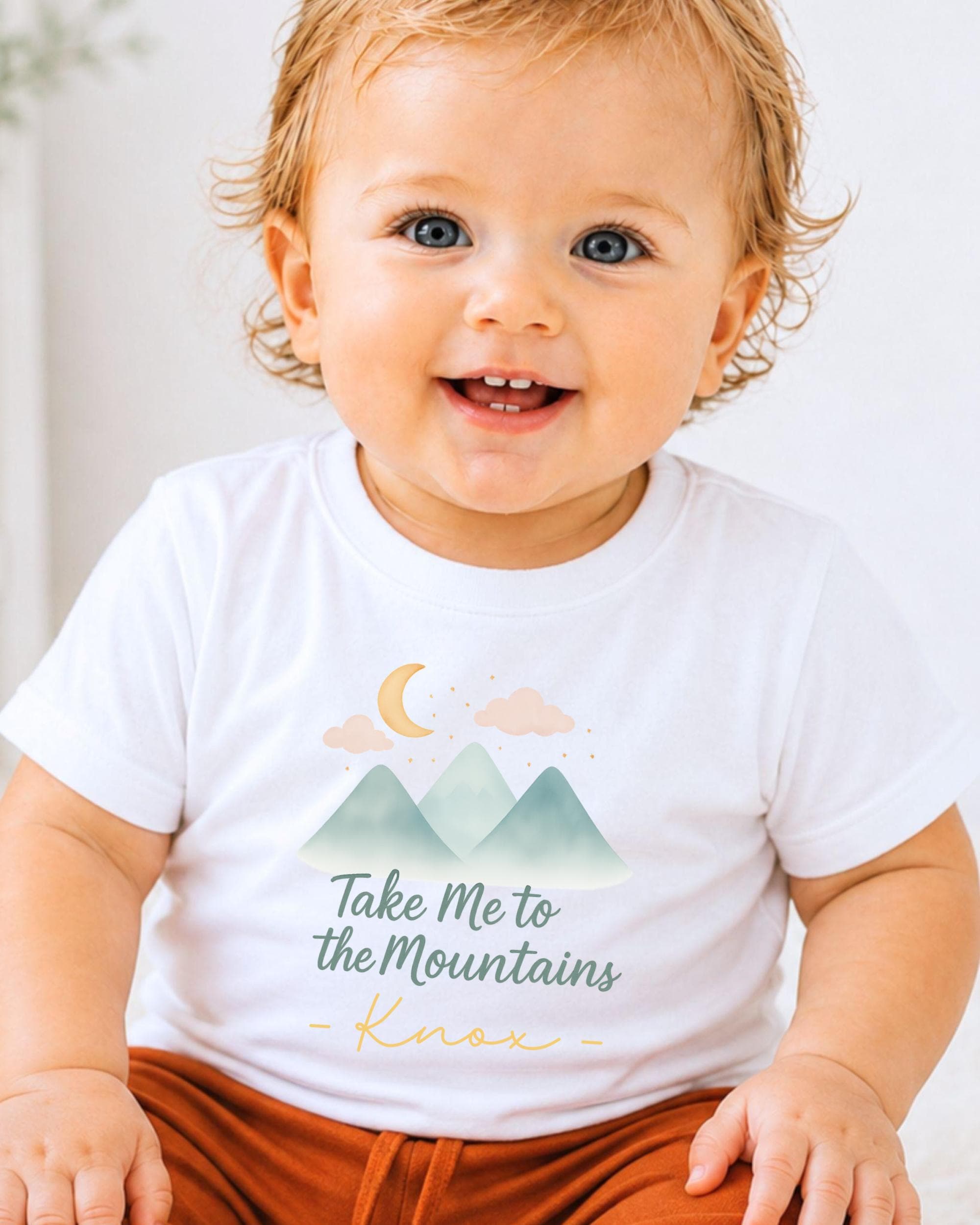 A smiling baby wearing a white t-shirt with a mountain and moon graphic and the text "Take Me to the Mountains - Knox" printed on it.