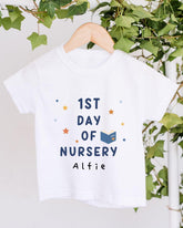 A white t-shirt with the text "1st DAY OF NURSERY" printed on it, along with a small illustration of a book and stars. The t-shirt is hanging on a wooden hanger, and there are green leaves visible in the background.