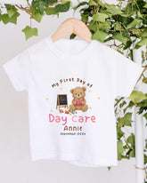A white t-shirt with a teddy bear and a chalkboard, along with the text "My First Day of Day Care Annie September 2023".