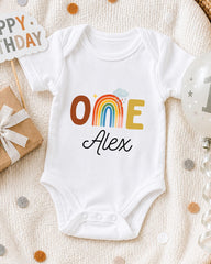 A white onesie with the text "ONE Alex" printed on it, surrounded by birthday decorations and a "Happy Birthday" sign.
