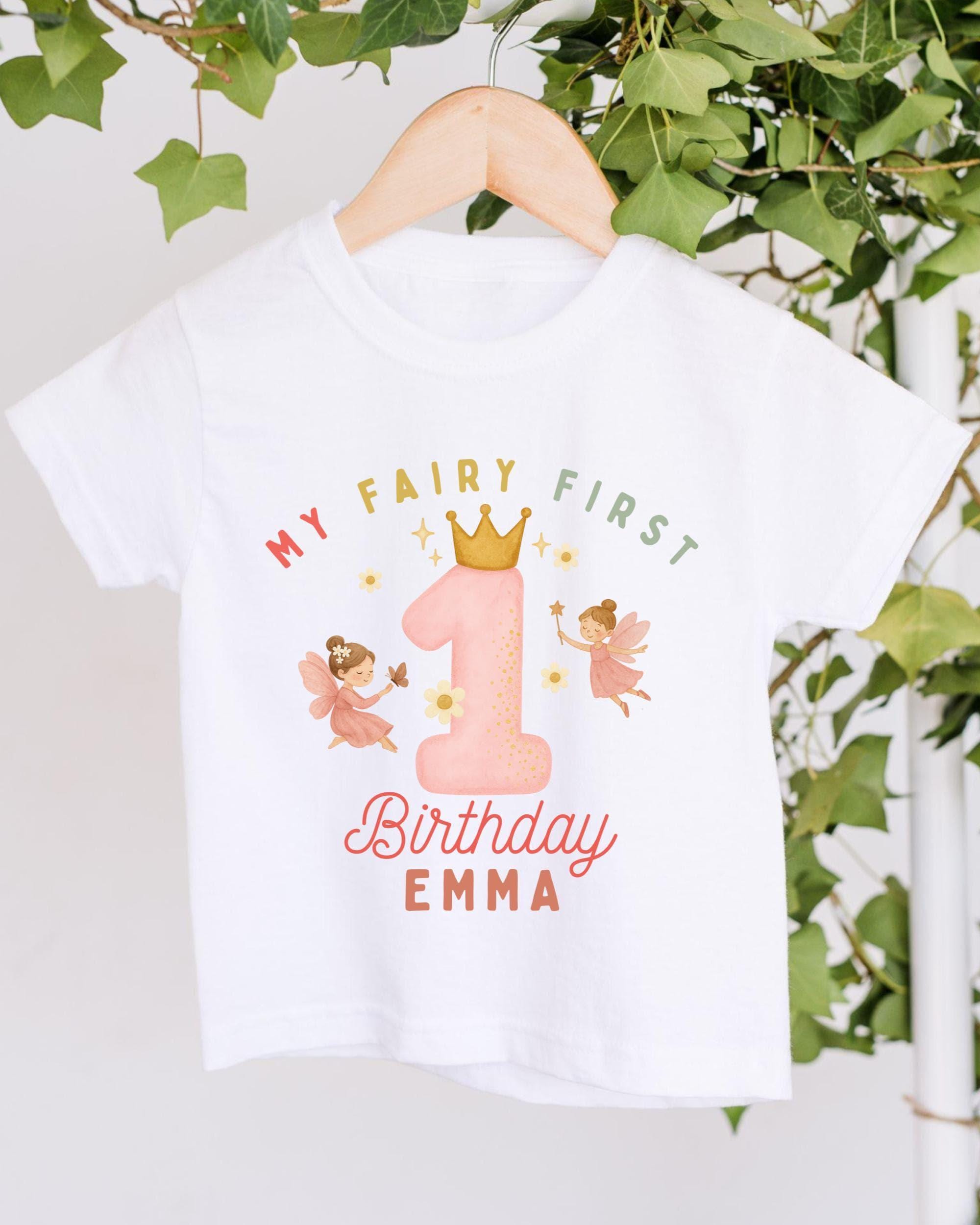 A white t-shirt with a graphic design featuring a pink number one and two-tailed fairies, along with the text "My Fairy First Birthday Emma" printed on it.