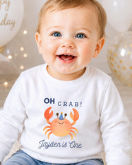 A smiling baby boy wearing a white shirt with a crab graphic and text that says "OH CRAB! Jayden is One".