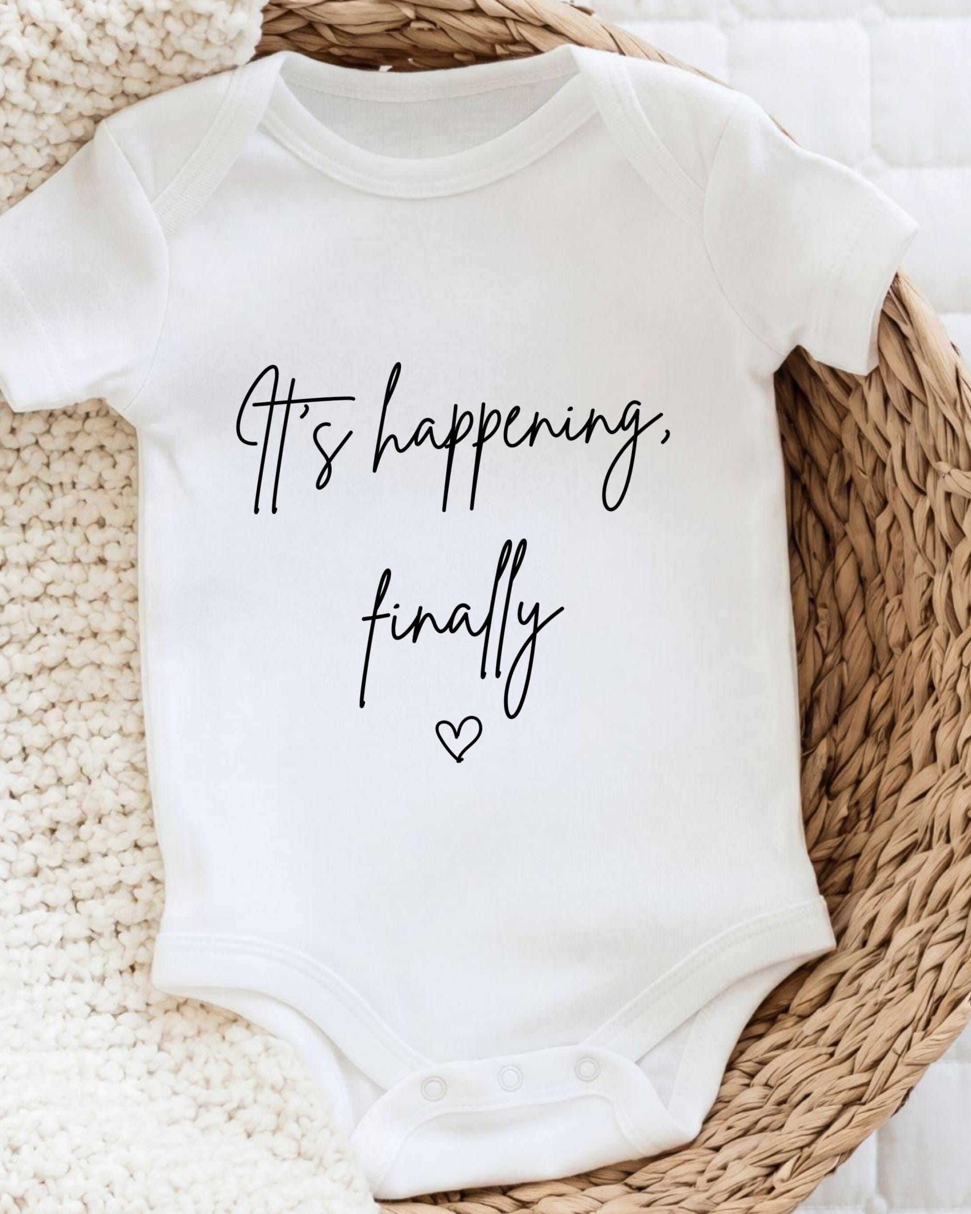 A white baby onesie with the text "It's happening, finally" printed on it, placed on a woven basket.