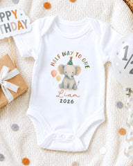 A white onesie with a cartoon elephant and the text "Half Way to One" printed on it, along with the name "Liam" and the year "2026". The onesie is placed on a white blanket, surrounded by birthday decorations such as a "Happy Birthday" sign, a balloon, and confetti.