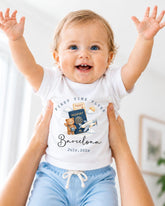 A smiling baby wearing a white t-shirt with a graphic of a passport and airplane, being held up by someone's arms.