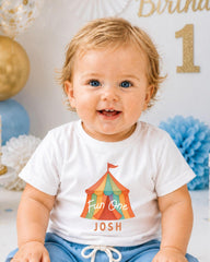 A smiling baby boy wearing a white t-shirt with a colorful circus tent graphic and the text "Fun One Josh" is sitting in front of a birthday party backdrop.