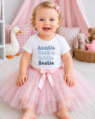 A smiling baby girl wearing a white t-shirt with the text "Auntie Tash's Little Bestie" and a pink tutu skirt, sitting on a white carpet surrounded by stuffed animals.