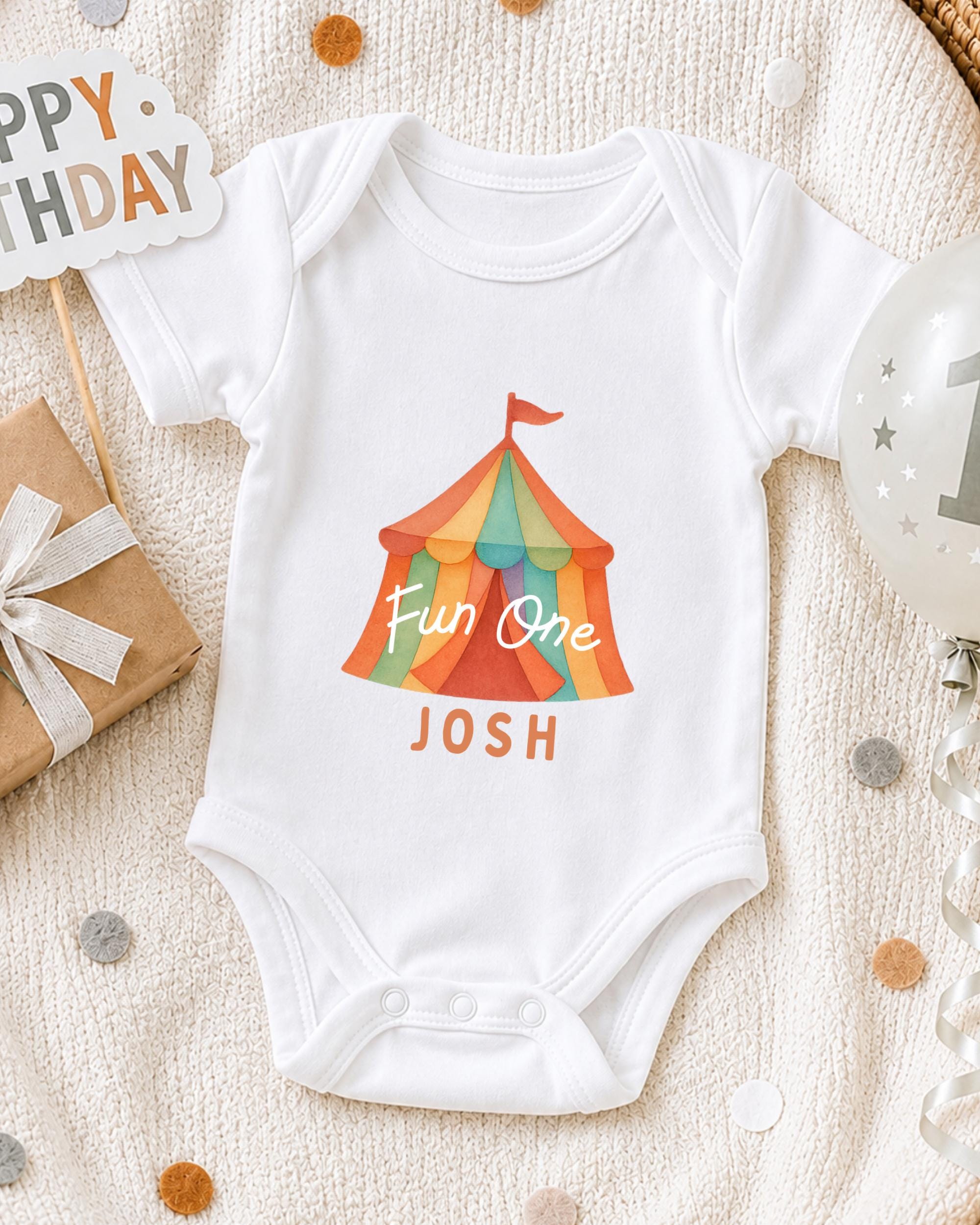 A white onesie with a colorful circus tent design and the text "Fun One Josh" printed on it, along with a "Happy Birthday" sign and a birthday balloon.