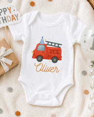 A white onesie with a red fire truck design and the name "Olivier" printed on it, along with a birthday sign and a balloon.