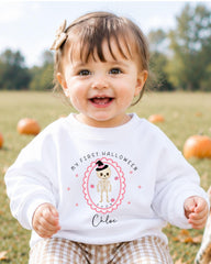 A smiling baby girl wearing a white sweater with a Halloween-themed design and text.