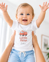 A smiling baby wearing a white t-shirt with a fire truck graphic and the text "Little Hero" is being held up by someone's arms.