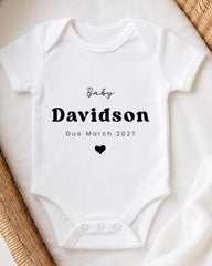 A white baby onesie with the text "Baby Davidson Due March 2027" printed on it.