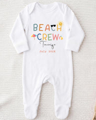 A white onesie with the text "BEACH CREW" printed on it, along with various beach-related icons such as a sun, sunglasses, and an umbrella.