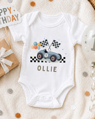 A white onesie with a race car design and checkered flags, along with a birthday sign and confetti, is displayed on a bed.