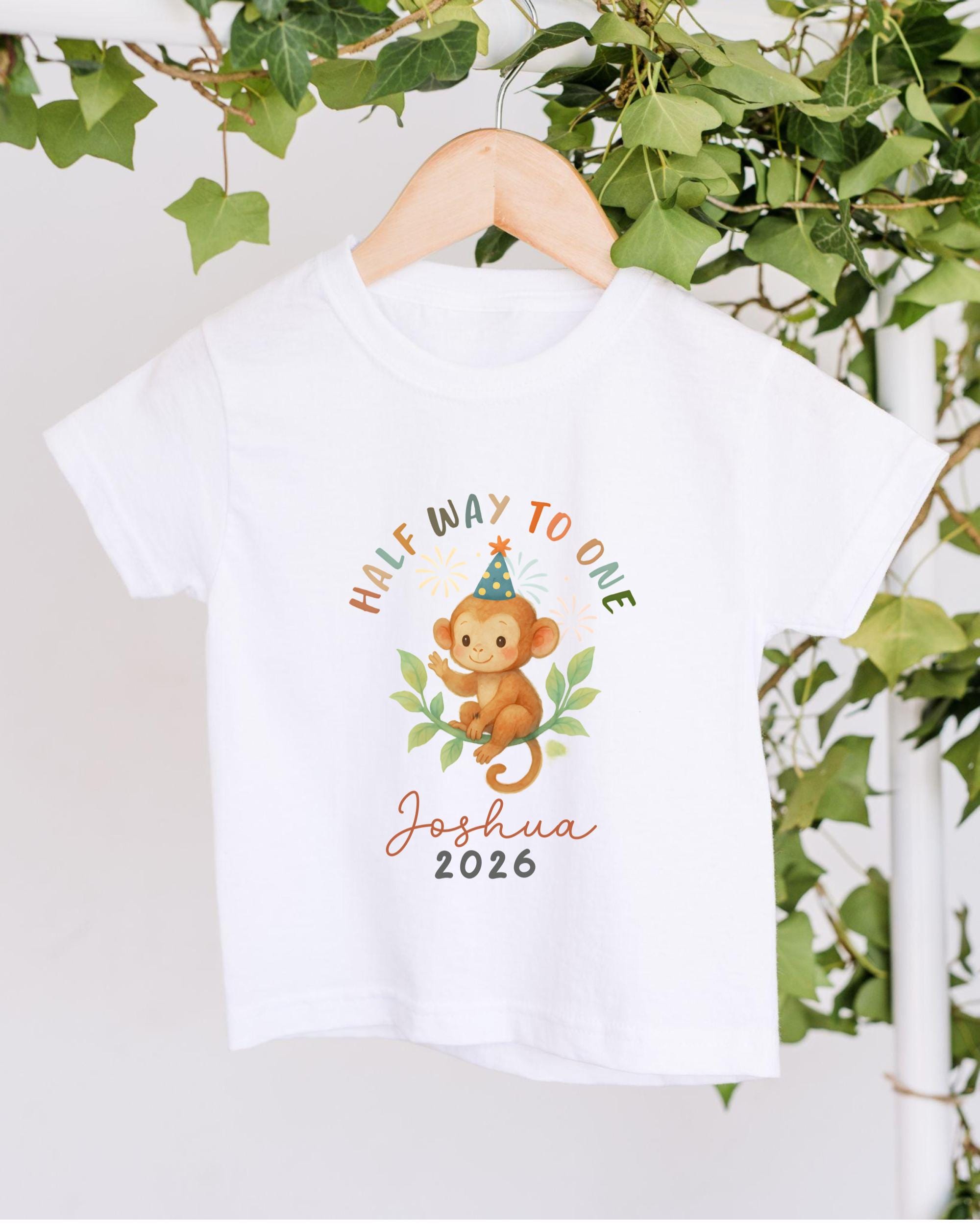 A white t-shirt with a cartoon monkey wearing a party hat and the text "Half Way to One Joshua 2016" printed on it, hanging on a wooden hanger against a backdrop of greenery.