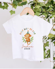 A white t-shirt with a cartoon monkey wearing a party hat and the text "Half Way to One Joshua 2016" printed on it, hanging on a wooden hanger against a backdrop of greenery.