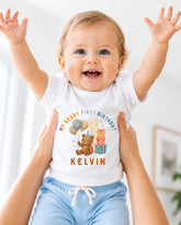 A smiling baby wearing a white t-shirt with a teddy bear and birthday decorations, being held up by someone's arms.