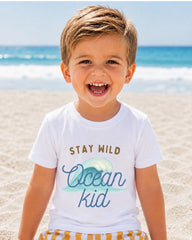 A young boy with blond hair is smiling and wearing a white t-shirt with the text "STAY WILD Ocean kid" printed on it, standing on a sandy beach with the ocean in the background.