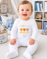A smiling baby boy wearing a white onesie with a graphic of three glasses and the text "Locally Brewed Jack - 2016" is sitting on the floor in a nursery setting.