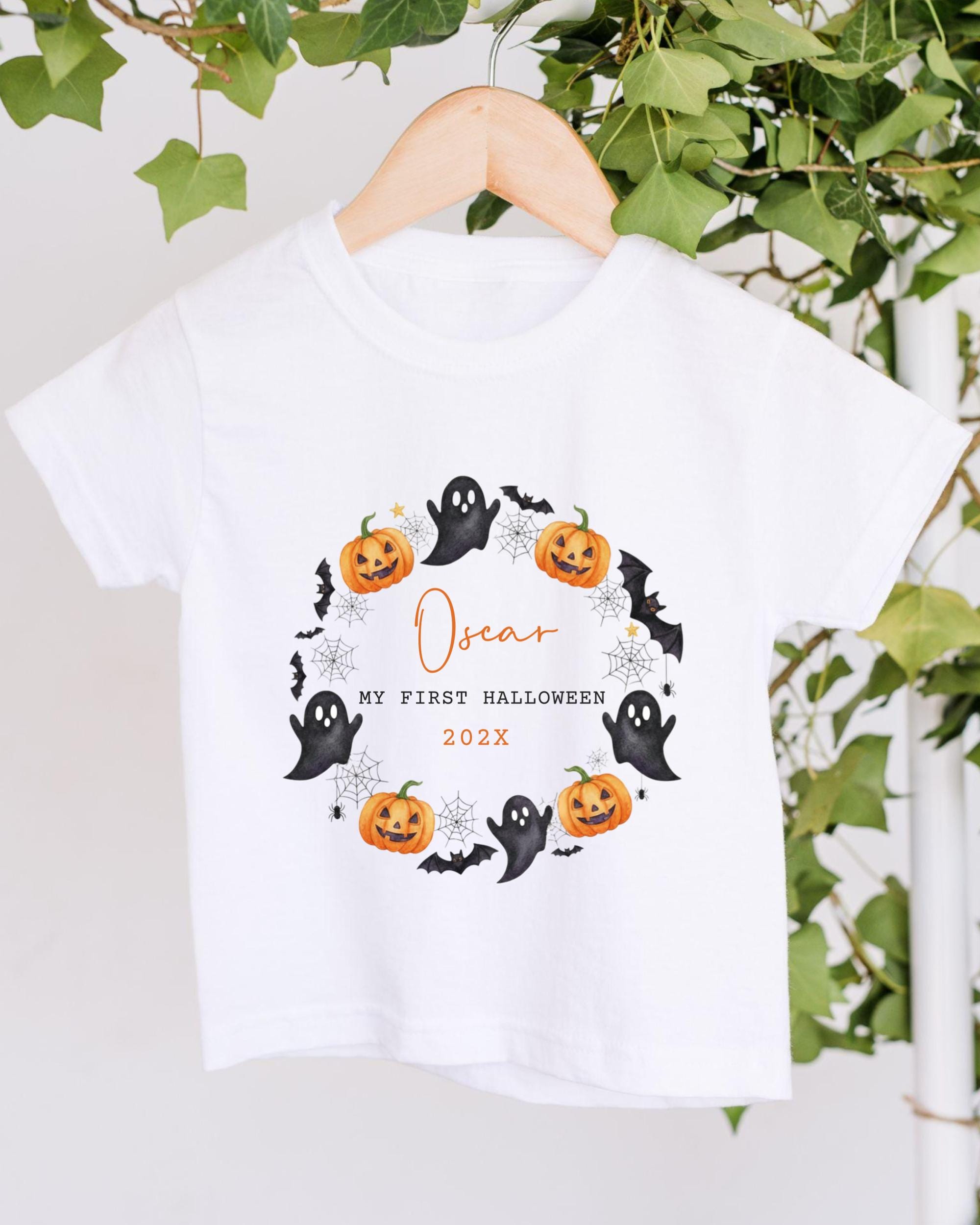 A white t-shirt with a Halloween-themed design hanging on a wooden hanger. The design includes a wreath of pumpkins, bats, and ghostly figures, along with the text "Oscar, my first Halloween 20x".