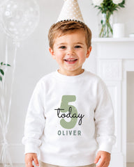 A young boy wearing a white sweatshirt with the number 5 and the text "Today Oliver" is smiling and posing for a photo.