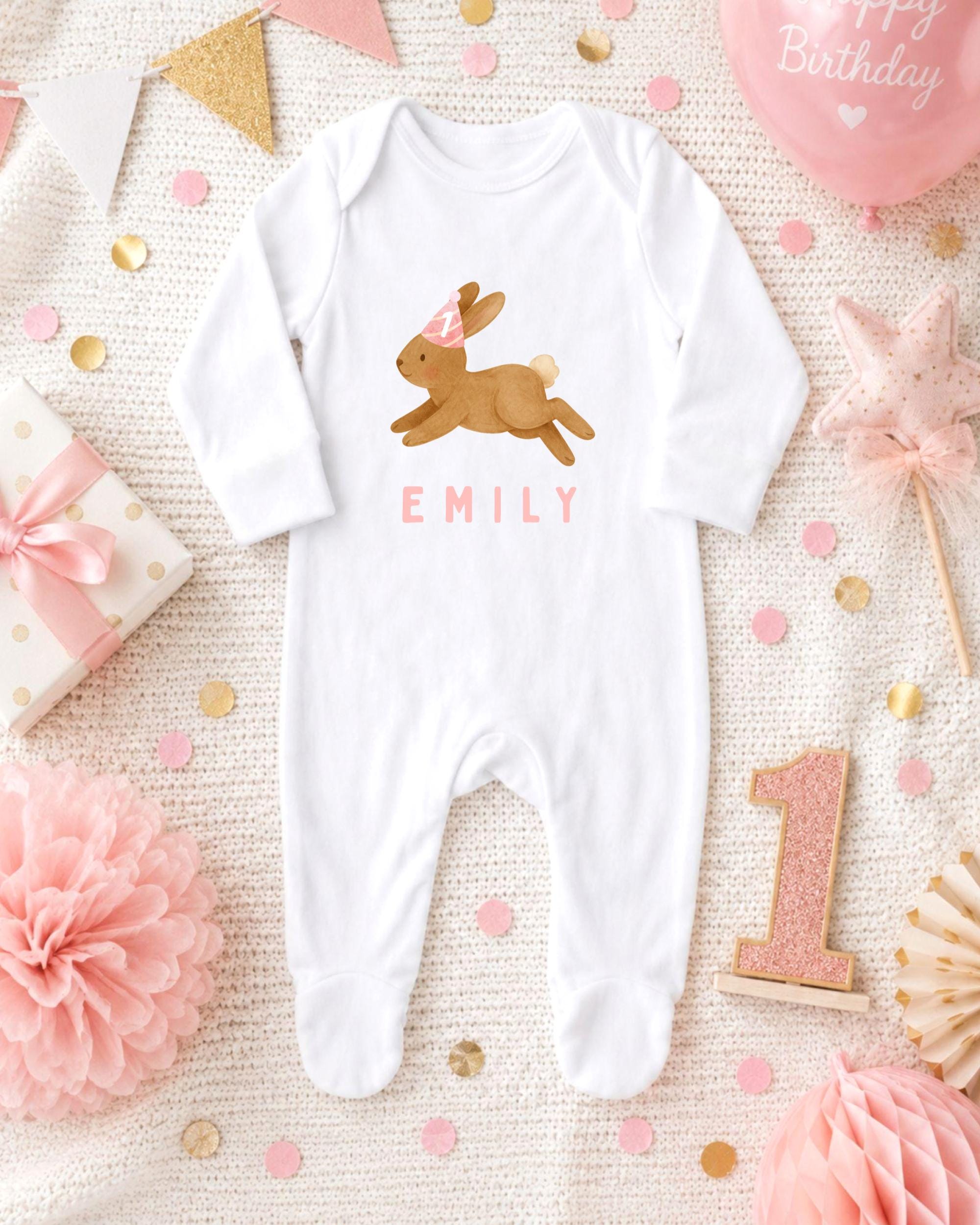 A white onesie with a bunny design and the name "EMILY" printed on it, along with a pink and gold birthday decoration, a pink star, and a pink and gold confetti arrangement.