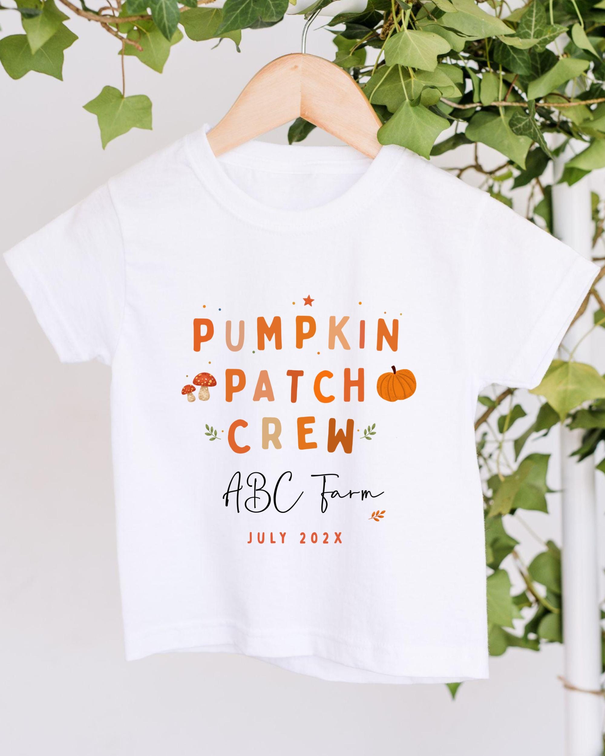 A white t-shirt with text and illustrations related to a pumpkin patch crew, hanging on a wooden hanger.