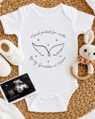 A white baby onesie with a hand-drawn wing design and the text "Hand picked for earth by my Grandma in Heaven" is displayed on a woven placemat. Next to the onesie are a crocheted bunny toy and a black and white photograph of an Ultrasound image of a baby's head.