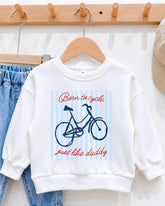 A white sweatshirt with a bicycle graphic and the text "Born to cycle just like daddy" printed on it, hanging on a wooden hanger.
