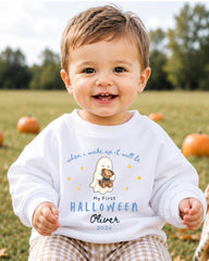 A smiling baby boy wearing a white sweatshirt with a graphic of a ghost and a teddy bear, sitting in a field with pumpkins in the background.