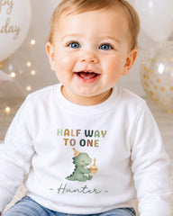 A smiling baby boy wearing a white shirt with a dinosaur graphic and text that says "Half Way to One Hunter".