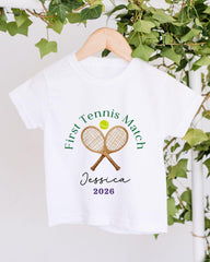 A white t-shirt with a tennis racket and ball graphic, along with the text "First Tennis Match" and " Jessica 2026" printed on it.